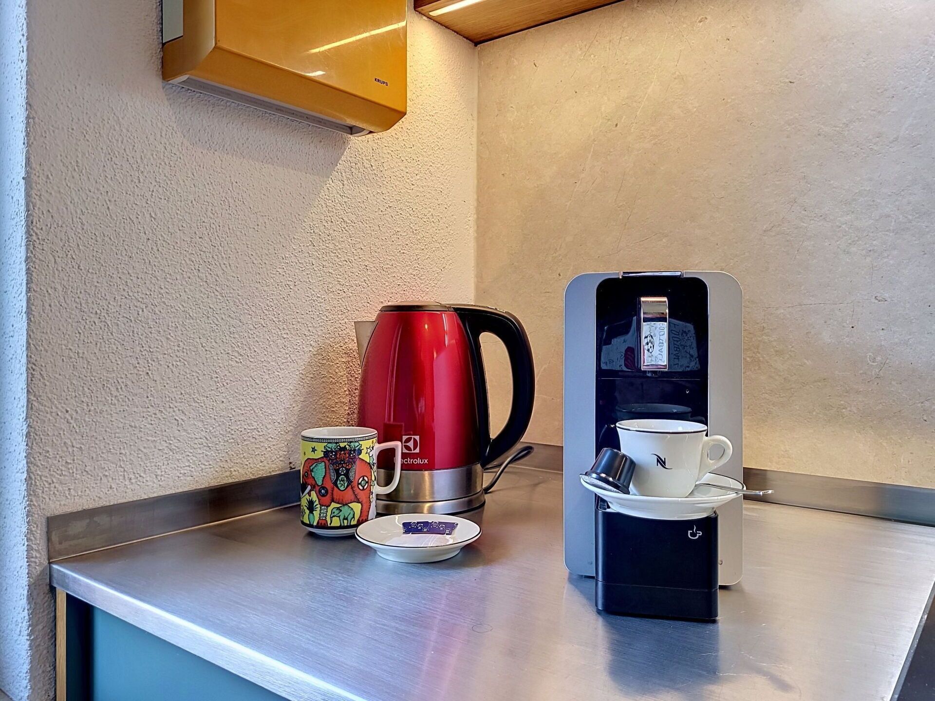 A stainless steel counter with a red coffee maker, a colorful mug, a white cup, and a small plate with tea bags.