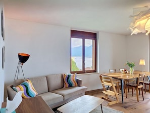 Ein modernes Wohnzimmer mit einer Couch, einem Esstisch mit Stühlen, einem großen Fenster mit Ausblick und einer Stehlampe.