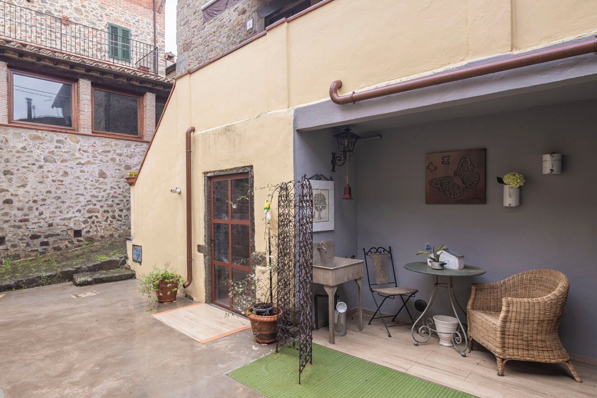 A courtyard with a stone wall, a door, a small table with chairs, and a wicker chair.