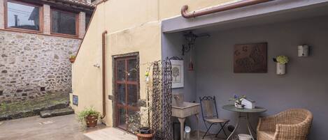 A courtyard with a stone wall, a door, a small table with chairs, and a wicker chair.