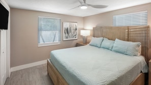 A bedroom with a bed, a nightstand, a lamp, a ceiling fan, and two windows with blinds. - Bare-Foot Bungalow (Sarasota)