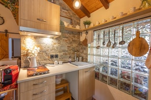 A kitchen with stone walls, wooden cabinets, a countertop with various kitchen items, and a window with hanging utensils.