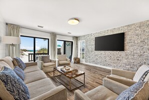 Ein modernes Wohnzimmer mit großem Fernseher, bequemen Sitzgelegenheiten und Blick auf den Strand.