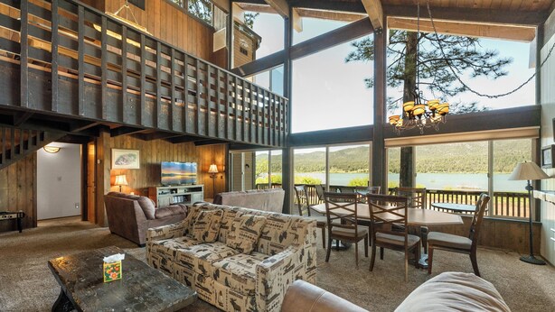 Un espacio habitable amplio con techo alto, vigas de madera, un gran ventanal con vistas a un lago, chimenea y zona de comedor con sillas y una mesa.