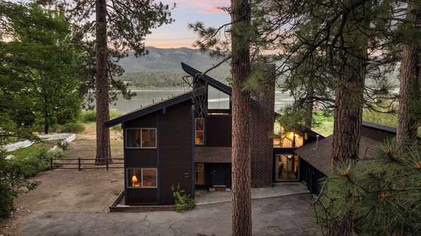 Una cabaña moderna de dos pisos con un amplio ventanal, rodeada de altos pinos, con vista a un lago.