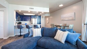 Un salon moderne avec un canapé d’angle bleu, un coin repas avec une table et des chaises, et une cuisine avec des armoires bleues et des appareils électroménagers en acier inoxydable.