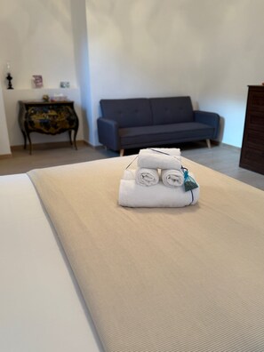 Un lit bien fait, agrémenté de serviettes pliées et d’un coussin décoratif, un canapé bleu foncé et une petite table en bois sur laquelle est posé un objet décoratif, le tout dans une pièce aux murs de couleur neutre.