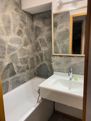 A bathroom with a stone wall, a white bathtub, and a sink with a mirror above it. - Sunny apartment just a 3-minute walk from the slopes (Chalon-sur-Saône)