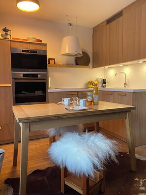 A modern kitchen with wooden cabinets, a built-in oven, a hanging light fixture, and a dining table with a fluffy white pouf. - Sunny apartment just a 3-minute walk from the slopes (Chalon-sur-Saône)