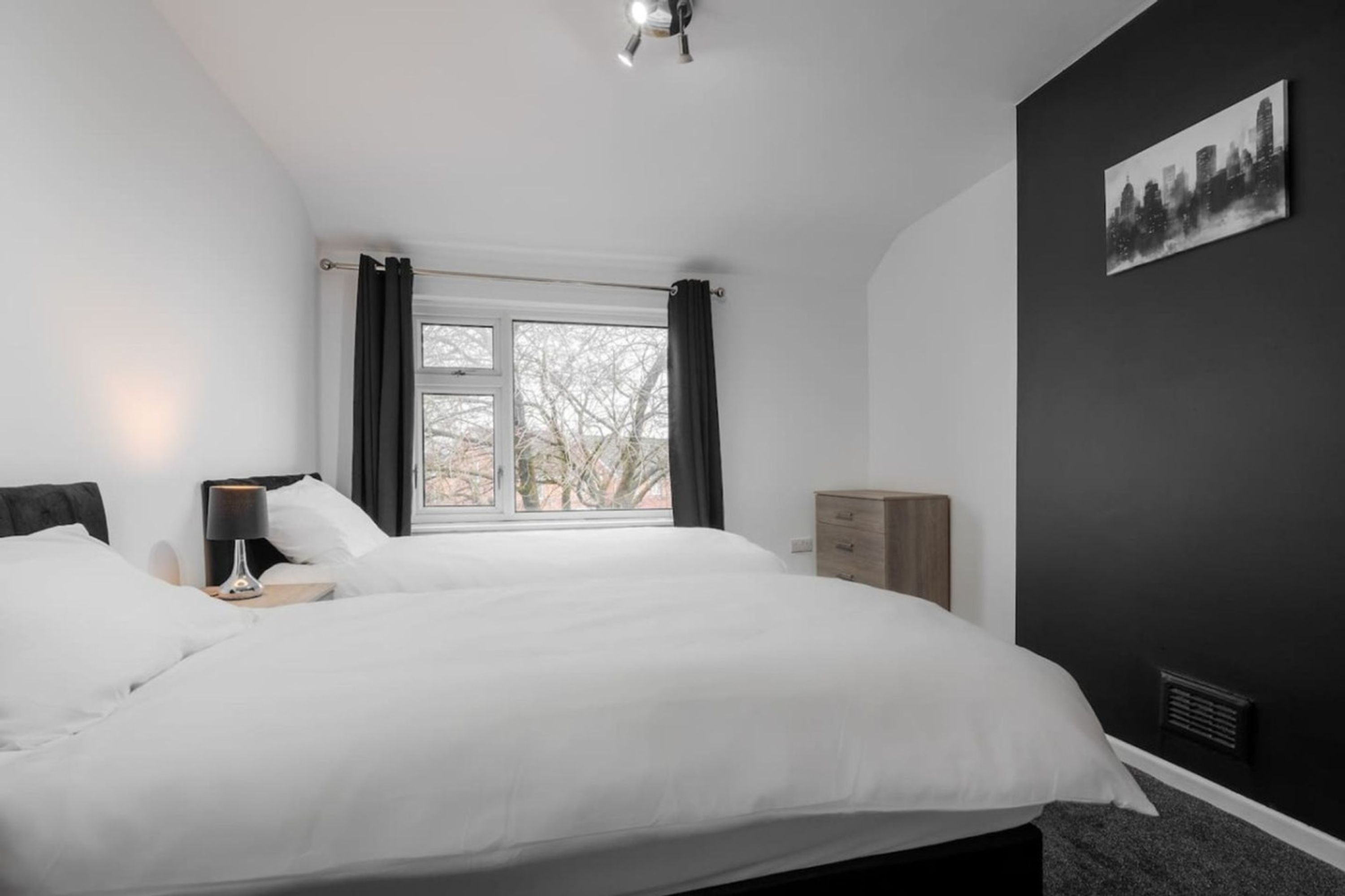 Une chambre avec un grand lit, une fenêtre avec des rideaux, une table de chevet avec une lampe, une commode et un tableau représentant un paysage urbain accroché au mur.