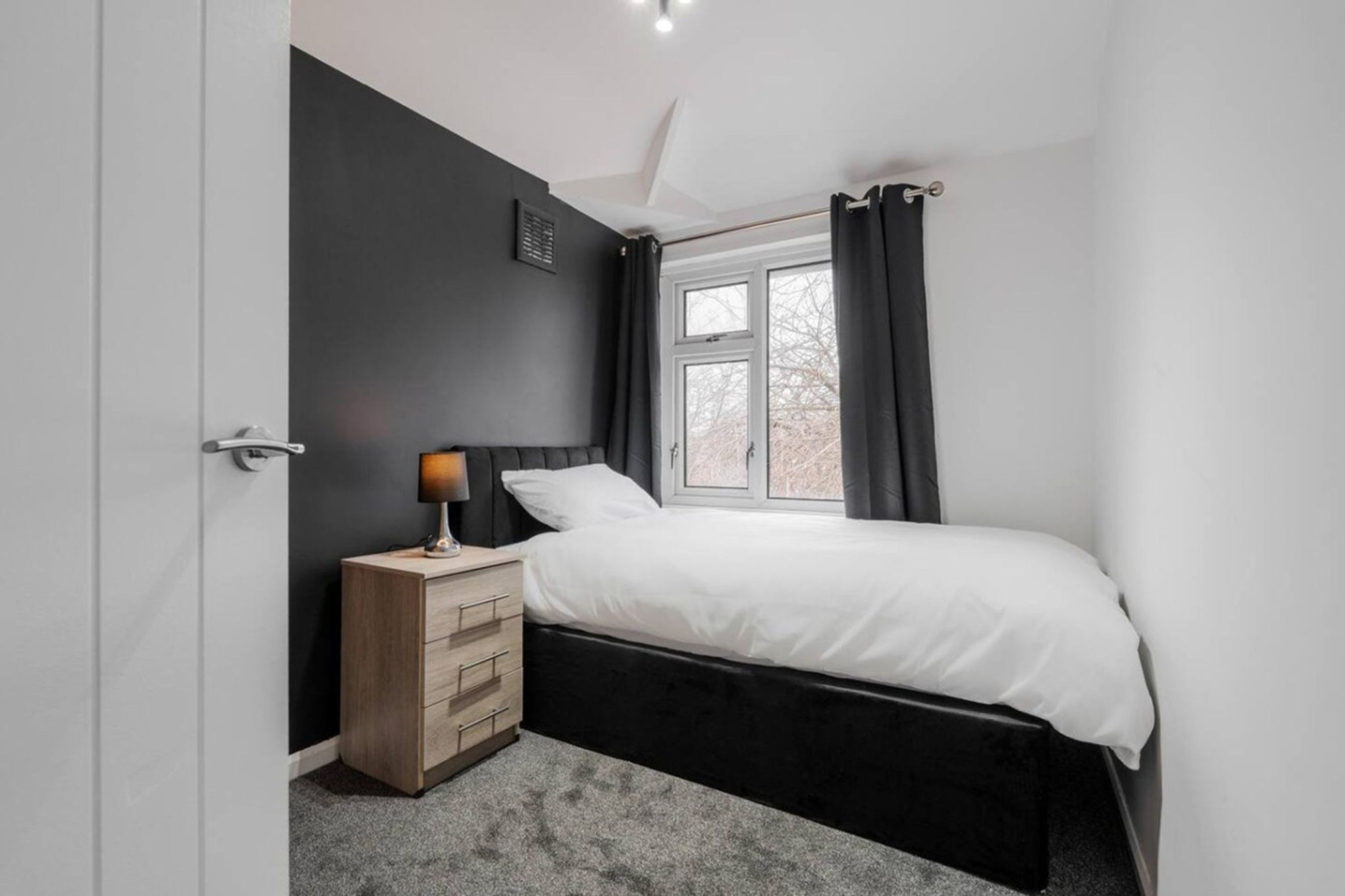 A bedroom with a bed, a nightstand, a lamp, a window with curtains, and a door.