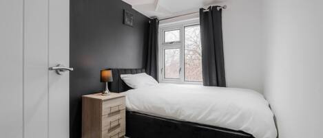 A bedroom with a bed, a nightstand, a lamp, a window with curtains, and a door.