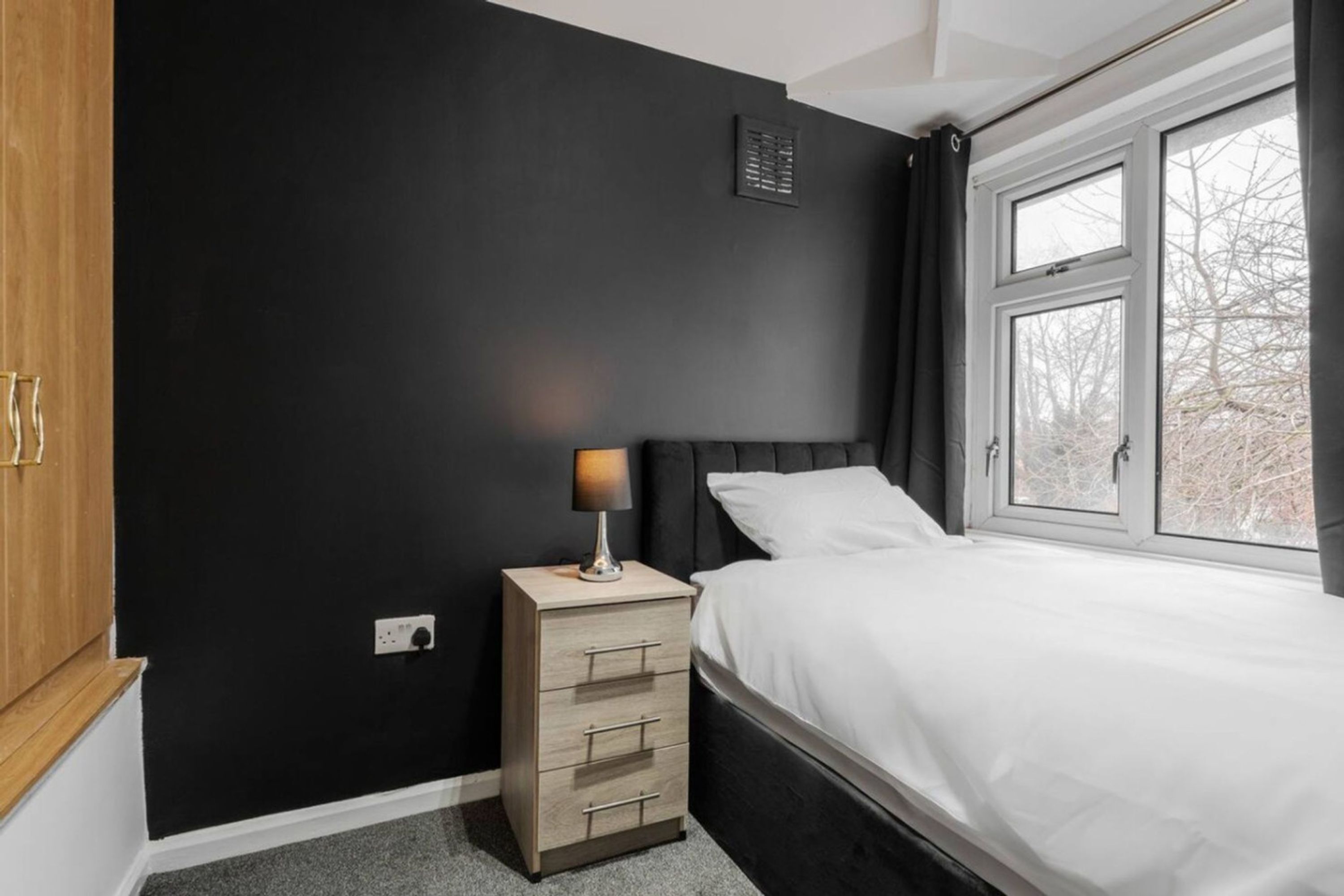 Et soveværelse med en seng, et natbord med lampe, et skab og et vindue med gardiner.