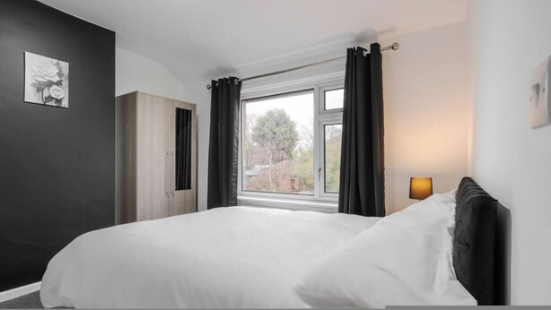 A bedroom with a bed, a window with curtains, a bedside lamp, and a painting on the wall. - Newly Refurbished 3BR Home Parking Sleeps 5 (Bolton)