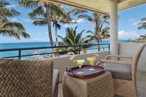 A balcony with wicker furniture, a tray with drinks, and a view of the ocean and palm trees.