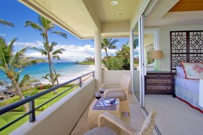 A balcony with a view of the ocean, a wicker chair, a small table with a glass of red wine, and a bed with a decorative headboard.