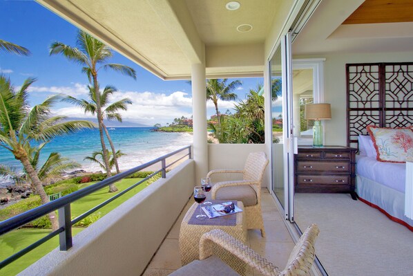A balcony with a view of the ocean, a wicker chair, a small table with a glass of red wine, and a bed with a decorative headboard.