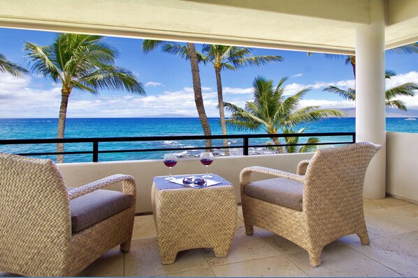A balcony with wicker furniture, two glasses of red wine, and a view of the ocean with palm trees.