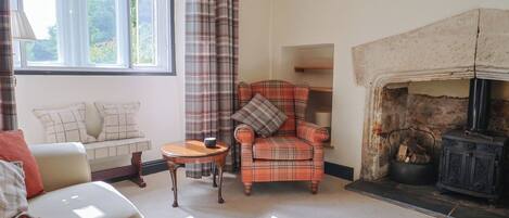 A cozy living room with a fireplace, a plaid armchair, a sofa, and a window with curtains.