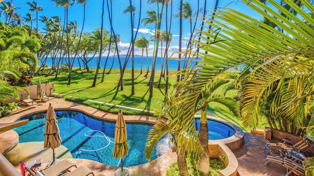 Ein Poolbereich mit Liegestühlen und Sonnenschirmen, umgeben von Palmen und Blick aufs Meer.