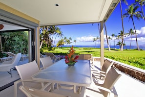 Eine Terrasse im Freien mit Tisch und Stühlen, einer Blumenarragemant und Blick auf das Meer und Palmen.