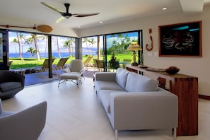 Ein modernes Wohnzimmer mit einer Couch, Sesseln, einem Deckenventilator und großen Fenstern mit Blick auf Palmen und das Meer.