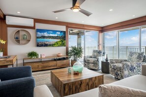 A modern living room with a wooden coffee table, blue and white patterned chairs, a large window with a view of the sea, and a ceiling fan. - Oceanfront- Supreme Setting (Lahaina)
