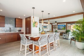 A modern kitchen with a central island, bar stools, pendant lighting, and a dining area with a view. - Oceanfront- Supreme Setting (Lahaina)
