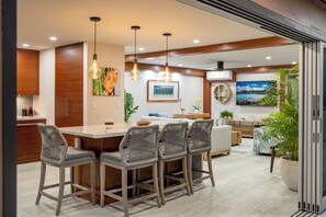 A modern kitchen with a central island, bar stools, pendant lighting, and a dining area with a sofa and coffee table. - Oceanfront- Supreme Setting (Lahaina)