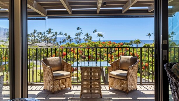 Un balcon agrémenté de fauteuils en osier et d’une table en verre, offrant une vue sur un jardin tropical et l’océan.
