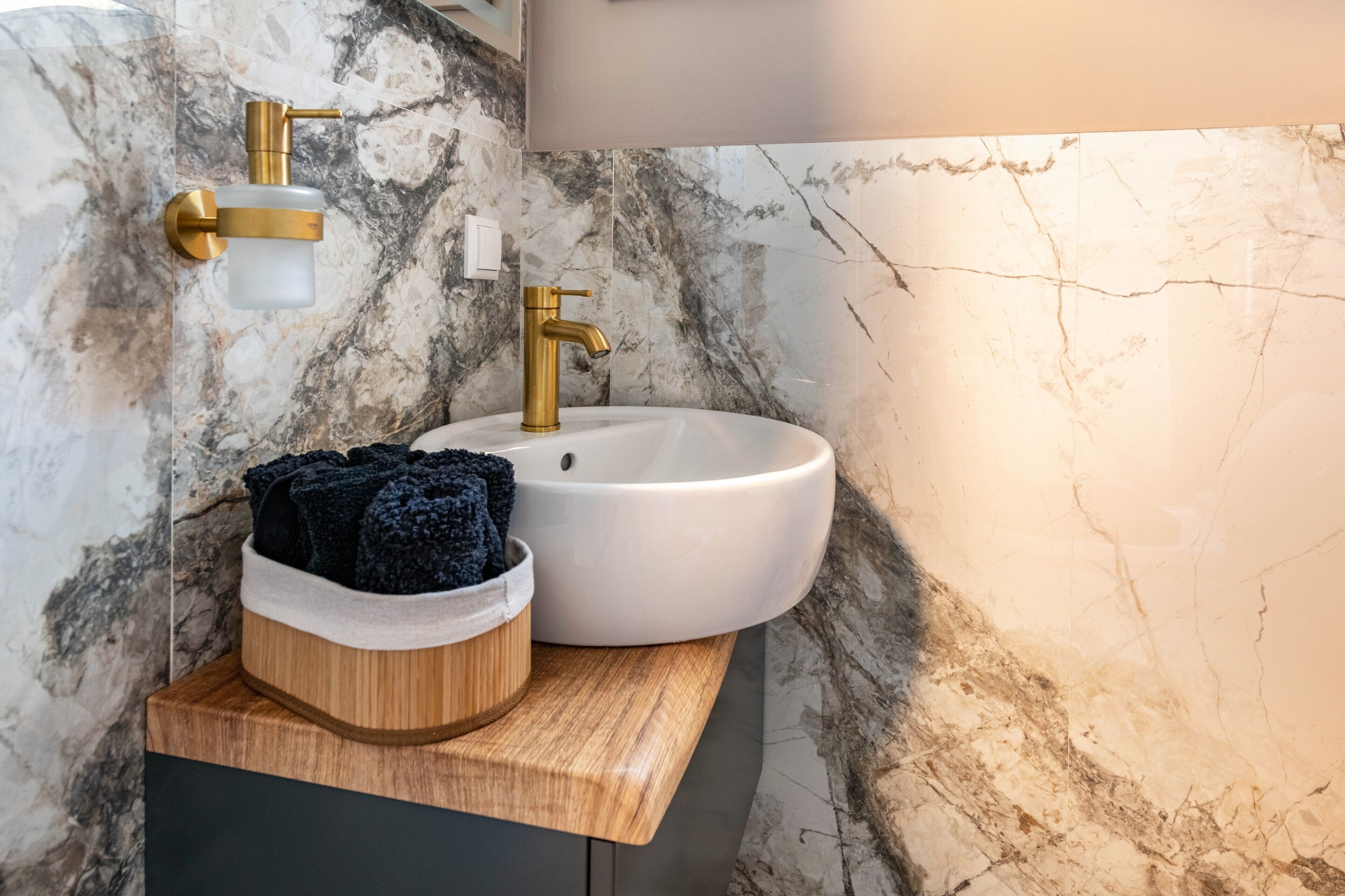 Un baño con pared de mármol, un lavamanos blanco, un grifo dorado y un mueble de tocador de madera.