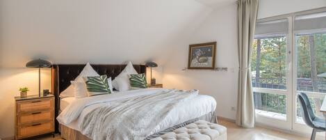 Un dormitorio con una cama grande, mesitas de noche, un banco, una ventana con cortinas y un cuadro enmarcado en la pared.