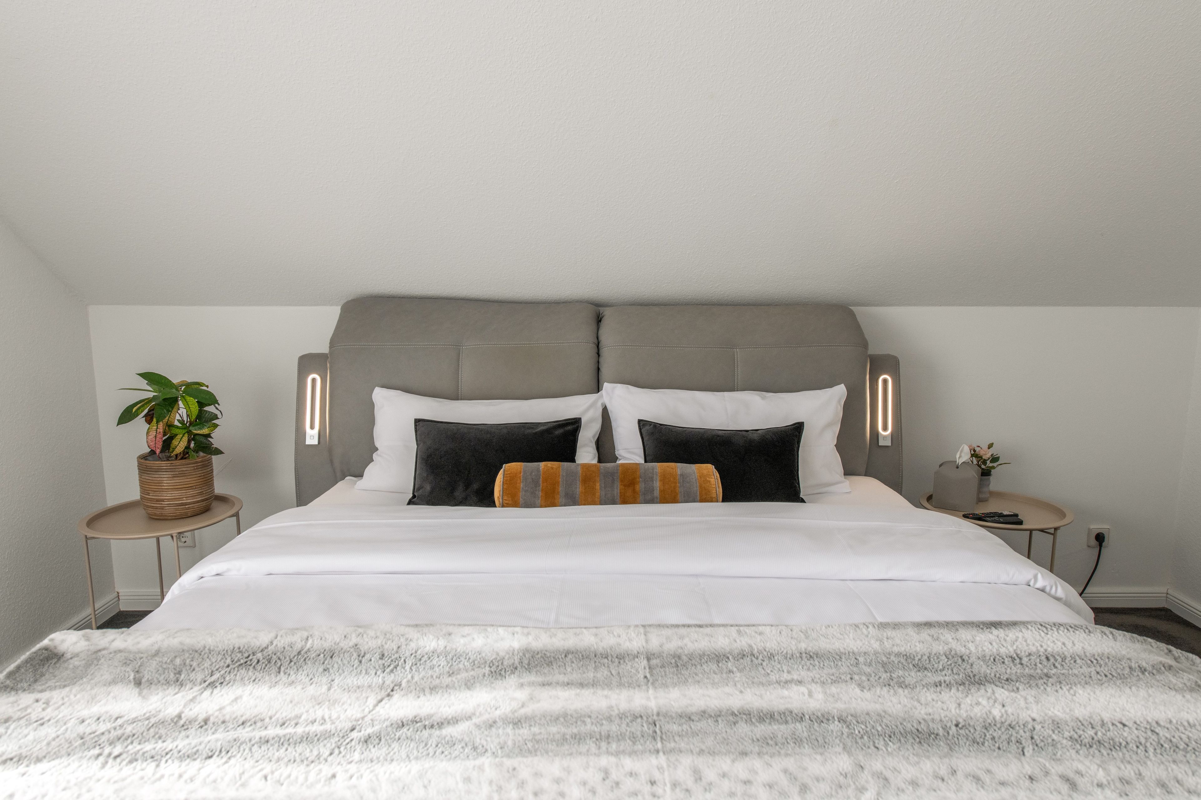Een net opgemaakt bed met een grijs bekleden hoofdbord, een potplant en een nachtkastje met een lamp.