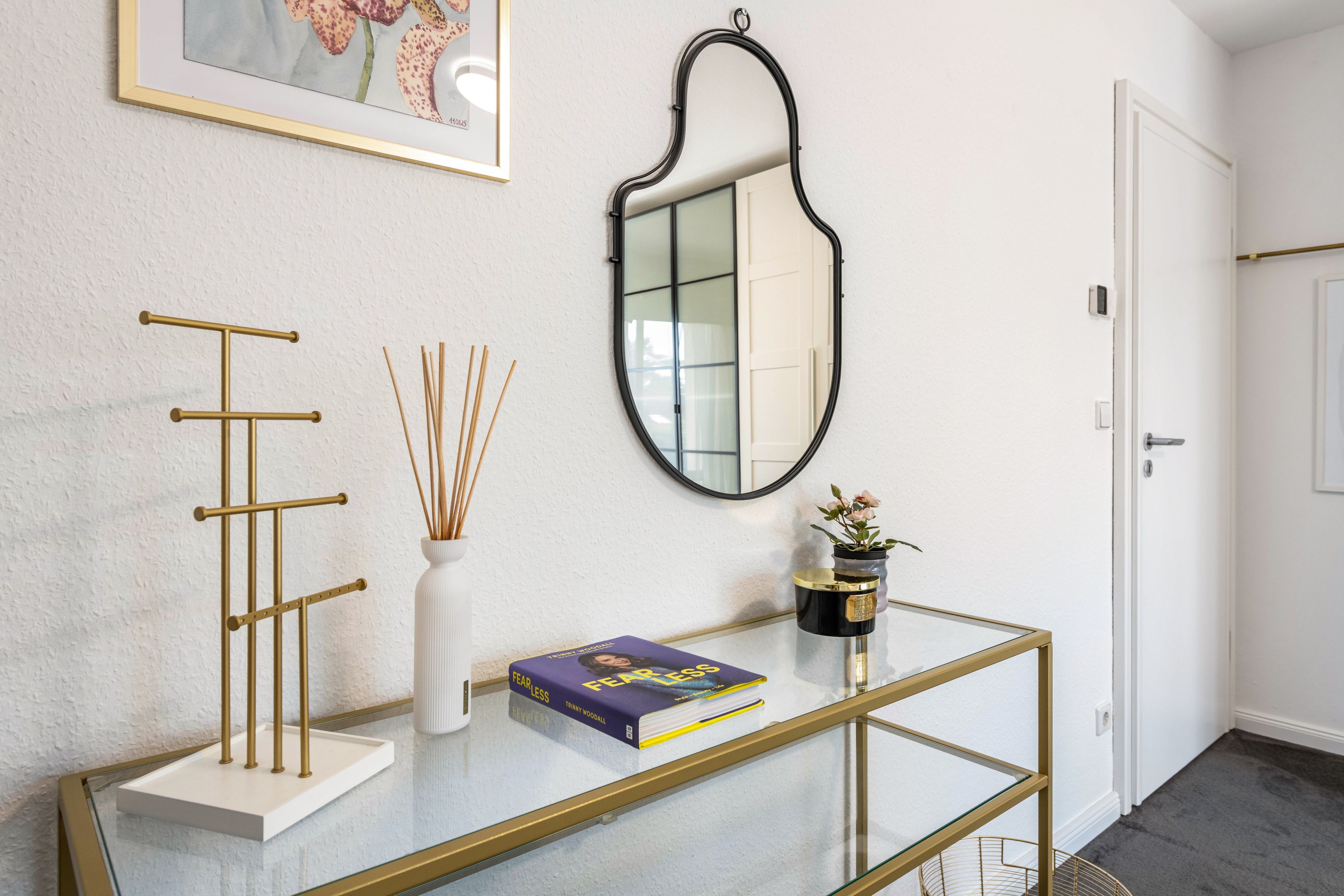 A hallway with a glass table, a gold stand, a framed picture, a mirror, and a door.