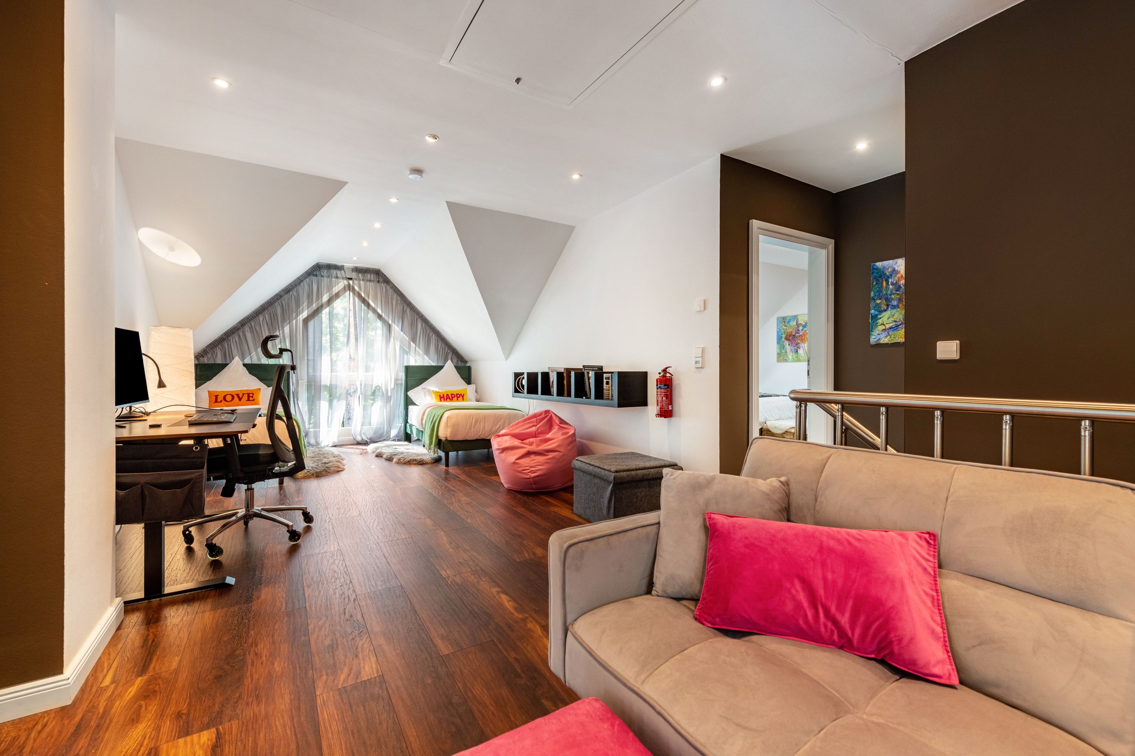 Un interior moderno con piso de madera, un sofá con un cojín rojo, un escritorio con una computadora, una cama con un letrero que dice 'HAPPY' y una escalera.