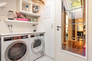 Un lavadero con lavadora y secadora de carga frontal, repisas con productos de limpieza y una puerta de vidrio que da a una sala de estar.