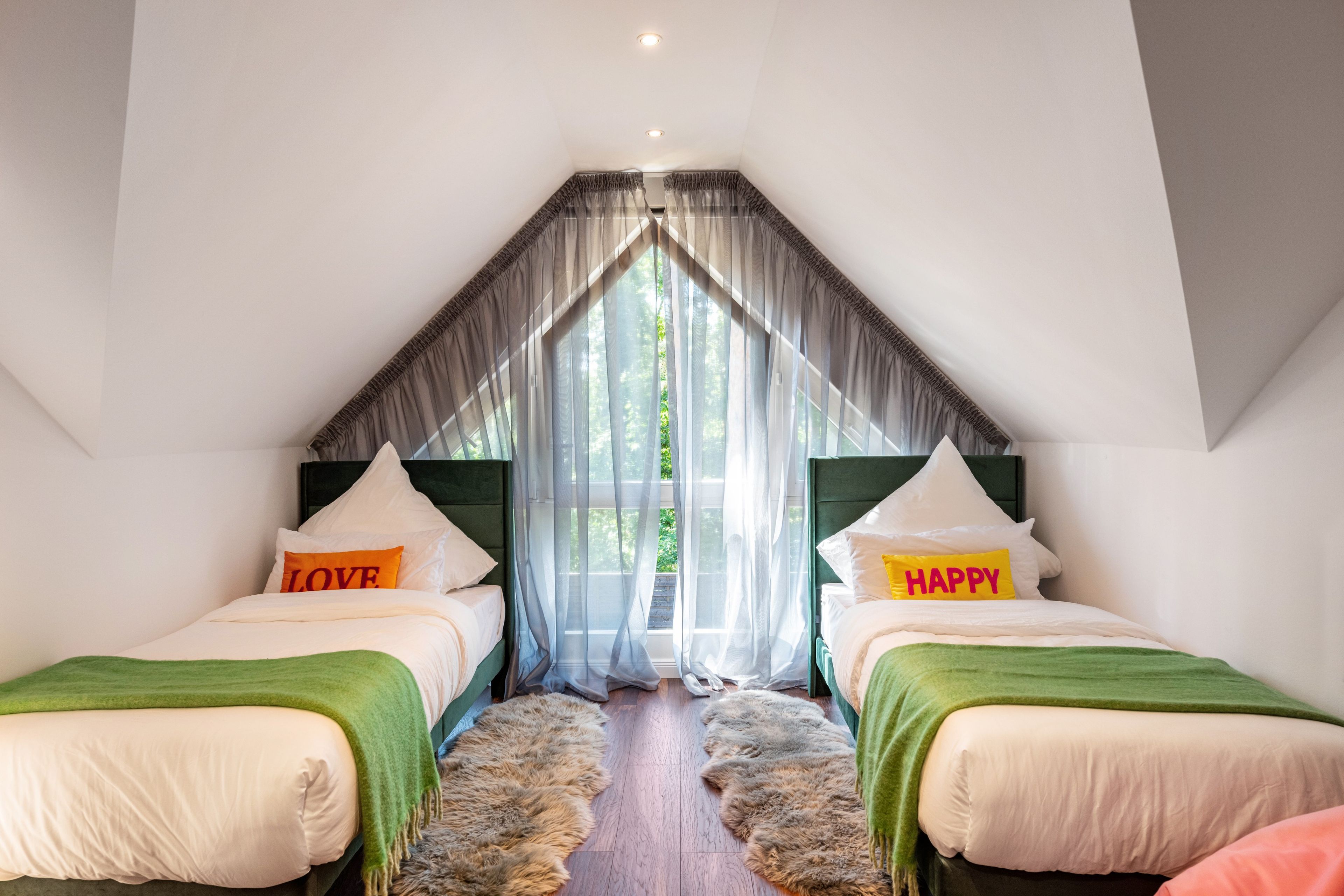 Une chambre avec deux lits, un plafond en pente, des rideaux légers et une vue sur la verdure.
