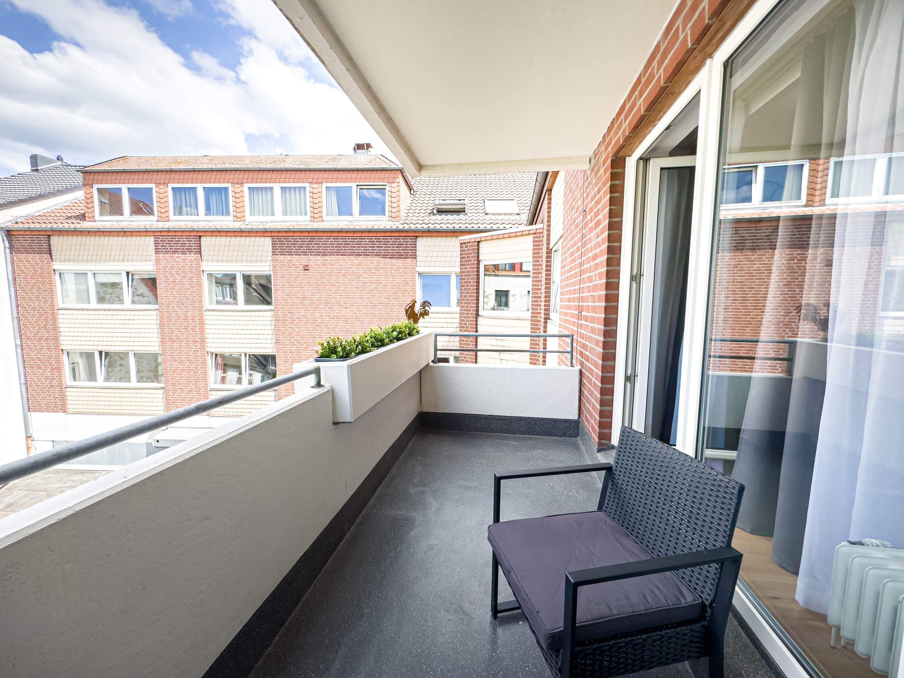 A balcony with a chair and a small plant, overlooking a residential area with brick buildings.