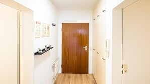 A narrow hallway with a wooden door at the end, a shelf with small plants, and a light switch on the wall. - Luchtbergstraße 1 (Bremen)