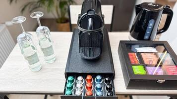 Una mesa con un parlante negro, dos botellas de vidrio, una cafetera negra y un cajón con bolígrafos de colores.
