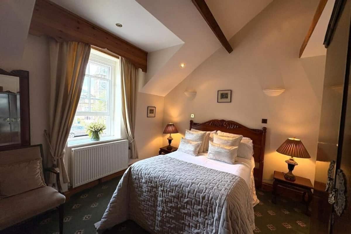 A bedroom with a bed, a window with curtains, a radiator, a wooden headboard, bedside lamps, and a chair.
