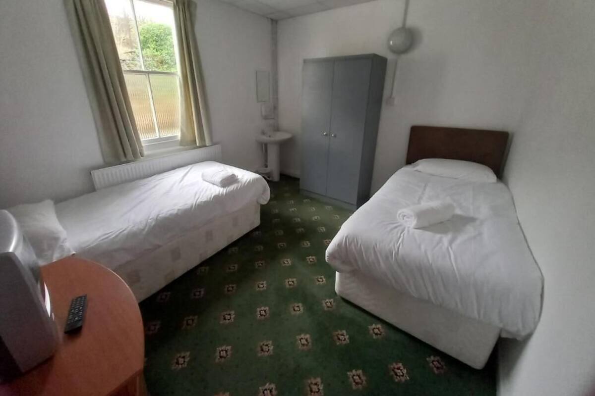 A hotel room with two single beds, a small round table, a window with curtains, a wardrobe, and a bedside lamp.
