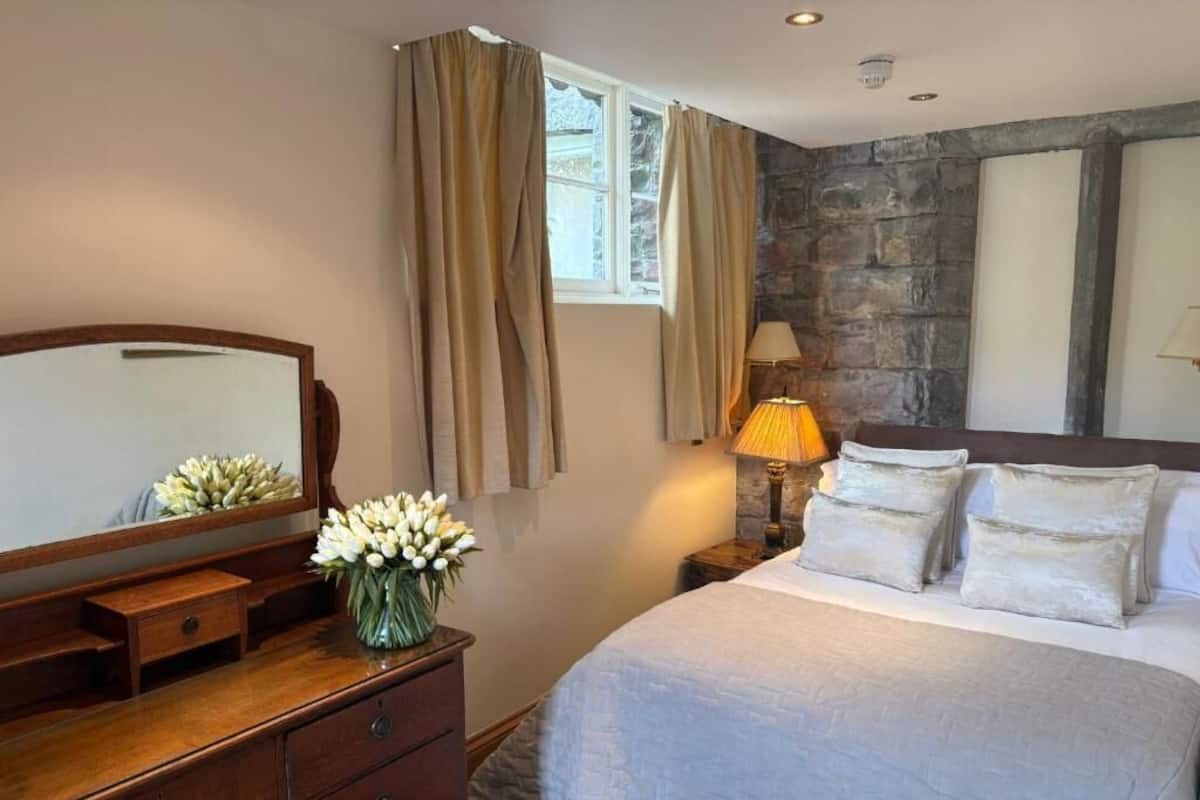 A bedroom with a bed, a dresser, a mirror, a window with curtains, a lamp, and a stone wall.