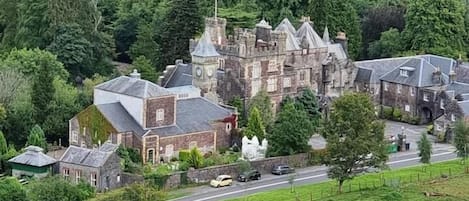 Un edificio de piedra grande e histórico con múltiples torres y una destacada torre del reloj, rodeado de abundante vegetación y una carretera en primer plano.
