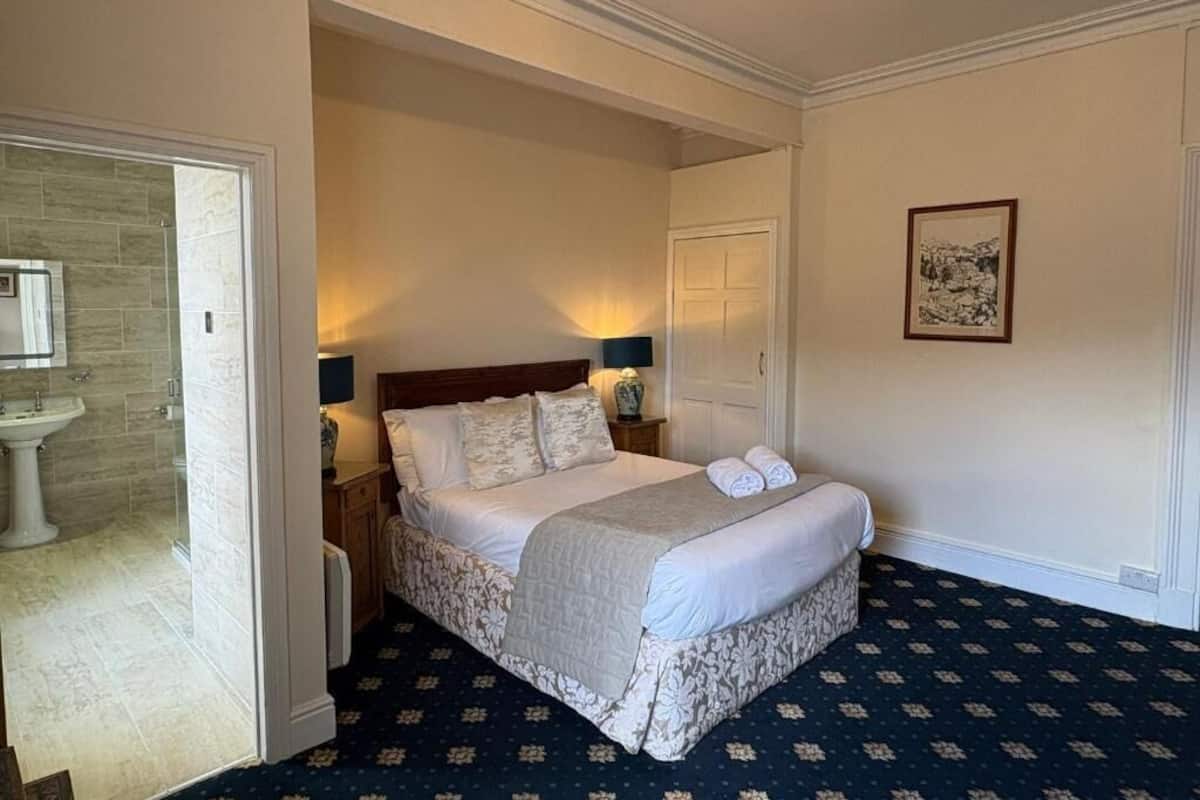 A hotel room with a bed, bedside lamps, a door, a framed picture, and a bathroom with a sink and mirror.