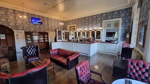 Un área de bar con mostrador y taburetes, un espacio de asientos con sillones de cuero y un televisor montado en la pared.