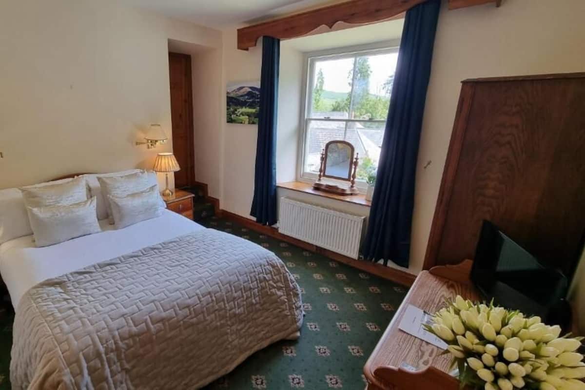 A bedroom with a bed, bedside lamps, a window with curtains, a radiator, a wooden desk with a television, and a bouquet of flowers.