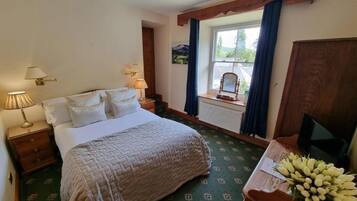 A bedroom with a bed, bedside lamps, a window with curtains, a radiator, a wooden desk with a television, and a bouquet of flowers.