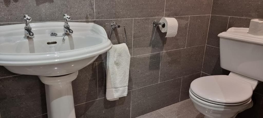 A bathroom with a white sink, two faucets, a towel rack with a white towel, a toilet, and a roll of toilet paper on the wall.