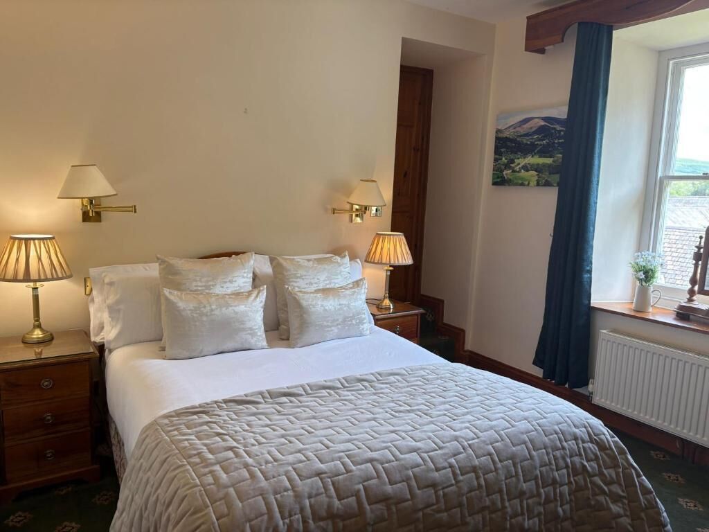 A neatly made bed with a quilt, two bedside lamps, a nightstand, a window with a view, and a painting on the wall.